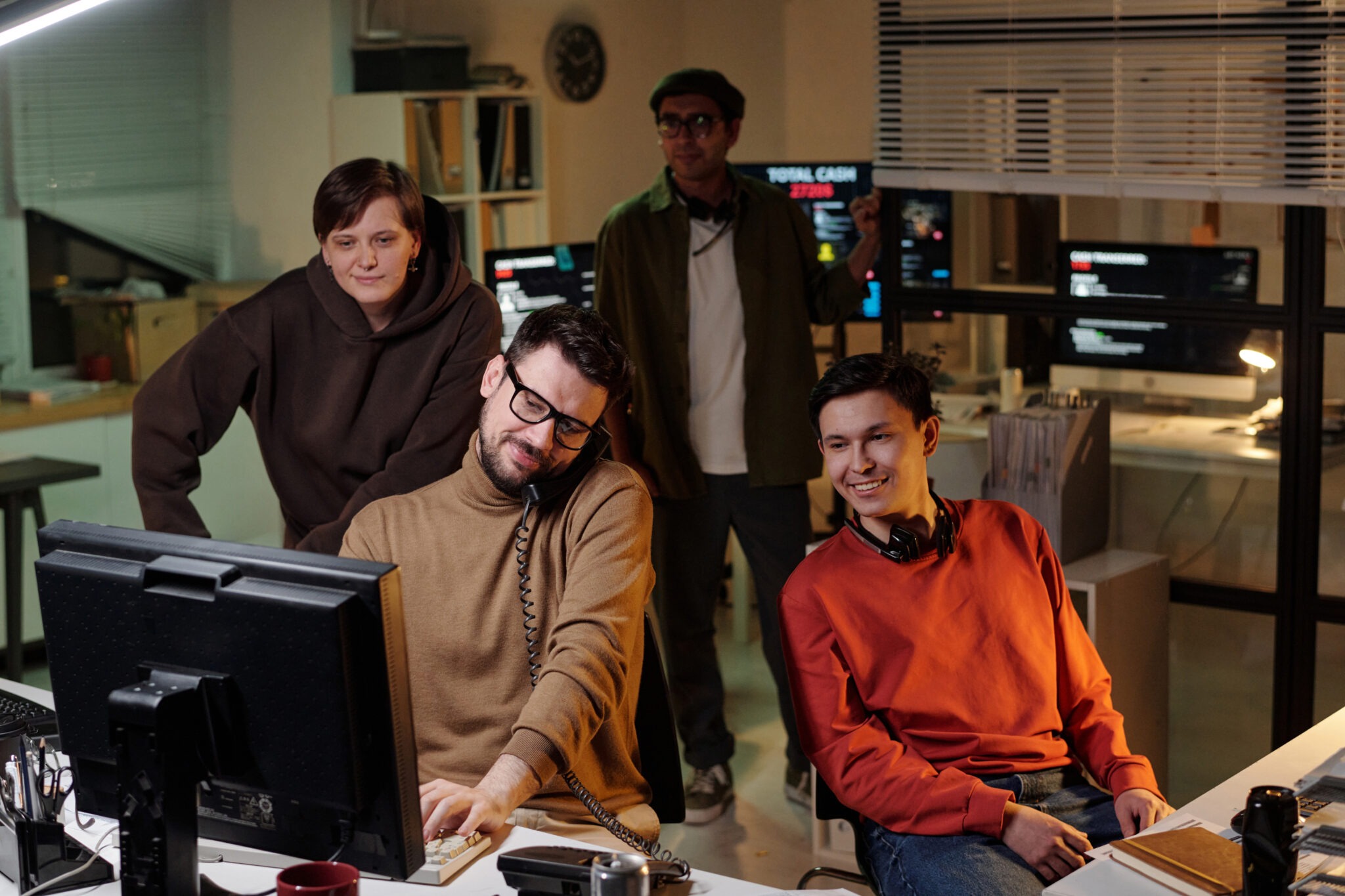 Group of people of diverse ethnicities operating computers and headsets in office at night, collaborating and monitoring screens